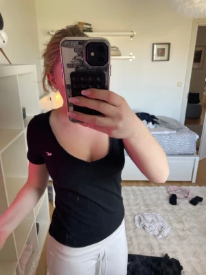 Svart v-ringad topp från Hollister - Svart v-ringad topp från Hollister med korta ärmar och figurnära passform. T-shirten har en liten logga på bröstet och är tillverkad i mjuk bomull, perfekt för en enkel och stilren look.