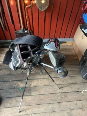 Golfset med bag, klubbor och tillbehör - Golfset med svart golfbag, flera klubbor inklusive putter och järn, samt tillbehör som golfhandske, greenlagare, markeringspenna och markeringsknapp. Perfekt för dig som vill komma igång med golf eller behöver extra utrustning. Märken som Callaway och FootJoy syns på utrustningen. Skriv om det är nåt som du är Intresserad AV