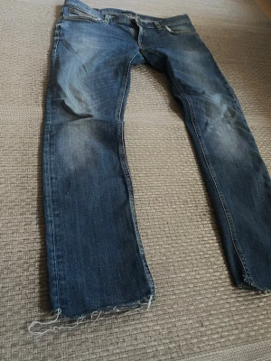 Blå raka Nudie Jeans med slitningar - Säljer ett par blå jeans med rak passform och slitna detaljer vid benslut och ficka. Jeansen har klassisk femficksdesign, normal midja och snygg tvätt med ljusare partier. Perfekta för en avslappnad och trendig look.
