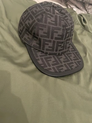 Fendi keps  - Snygg svart keps från Fendi med ikoniskt FF-mönster över hela kepsen. Böjd skärm med svart kant i läderlook och justerbar rem baktill. Perfekt accessoar för att lyfta din streetwear-stil.