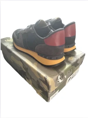 Valentino sneakers med camo-detaljer - Snygga sneakers från Valentino med svart och mörkgrön mocka, röda detaljer på hälen och gul sula. Skorna har coola nitar på bakkappan och kommer i en camo-mönstrad originalkartong. Perfekta för dig som gillar streetstyle och exklusiva märken. Sitter lite mindre i storlek 