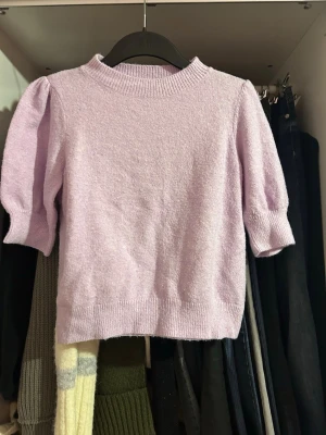 Ljuslila stickad topp Vero Moda - Supersöt ljuslila stickad topp från Vero Moda med puffärmar och ribbade kanter. Tröjan har en rund halsringning och är gjord i ett mjukt material som känns skönt mot huden. Perfekt för dig som gillar pastellfärger och trendiga detaljer.