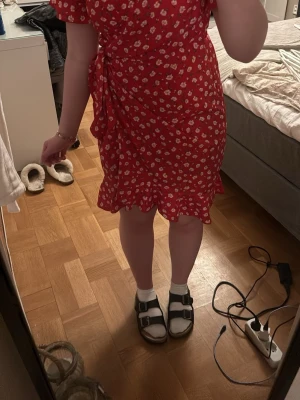 Röd blommig omlottklänning - Söt röd omlottklänning med vitt blommönster och volangkant. Klänningen har korta ärmar och knyts i midjan.