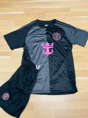 Svart och grå fotbollströja med shorts - Säljer en svart och grå fotbollströja med matchande shorts. Tröjan har ett rosa logotryck på bröstet och klubbmärke på vänster sida. Materialet är glansigt och lätt, perfekt för träning eller match. Shorts med elastisk midja och klubbmärke på benet.