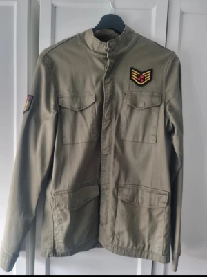 Olivgrön overshirt med patchar - Snygg olivgrön overshirt med militärinspirerade patchar på bröstet och ena ärmen. Jackan har fyra fickor framtill och en hög krage med knappstängning. 