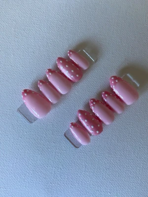 Rosa press-on naglar med prickar - Snygga press-on naglar i en mjuk rosa färg med vita prickar på toppen. Naglarna har en mandelformad design och är gjorda i plast. Perfekt för dig som vill ha en söt och trendig look utan att behöva gå till salong.