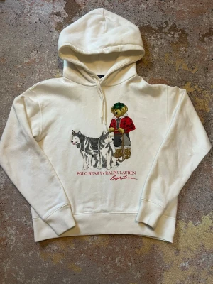 Vit hoodie Polo Bear Ralph Lauren - Vit hoodie från Ralph Lauren med Polo Bear-motiv framtill där en björn i röd jacka står med två huskies. Klassisk huva med dragsko och ribbade muddar. Tryckt text 'POLO BEAR by RALPH LAUREN' under motivet. Perfekt för en chill och stilren look.  Hoodien är använd max 5 gånger och är fortfarande mjuk och skön inuti. Är i storlek XS. Kontakta gärna vid frågor, jag postar så snabbt jag kan!