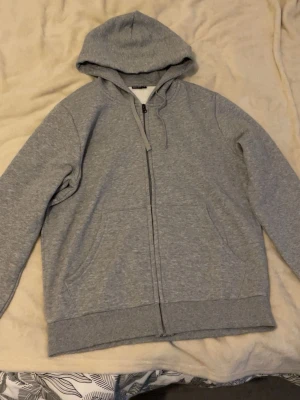 Grå zip-hoodie  - Klassisk grå hoodie med dragkedja från Take Off. Mjuk insida, huva med dragsko och två stora fickor framtill. Perfekt för chill dagar eller när du vill ha en enkel och stilren look. Materialet är bomullsblandning för extra komfort.