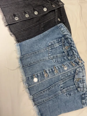 Två jeanstoppar med knappar - Säljer två snygga jeanstoppar en i ljusblå denim och en i svart. Båda har fransig nederkant, dekorativa silverknappar framtill och fickor. Perfekta för dig som gillar en trendig och avslappnad stil.