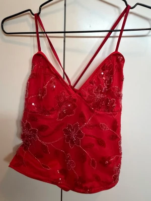 Röd blommig paljett linnetopp - Snygg röd linnetopp med tunna axelband och v-ringning. Toppen har blommigt broderi och paljetter över hela, samt knytning i ryggen för extra detalj. Perfekt för dig som vill sticka ut med en färgstark och unik look.