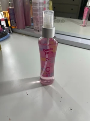 So...? Candy Floss Body Mist - Fräsch body mist från So...? i doften Candy Floss. Flaskan är genomskinlig med rosa vätska och har ett spraymunstycke i plast. Designen är söt med pastellfärgade ränder och text i rosa och lila. Perfekt för dig som gillar söta och lekfulla dofter.