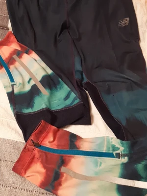 Färgglada leggings från New Balance - Snygga leggings från New Balance med coolt färgblockmönster i svart, turkos, grönt och rött. De har dragkedjor vid bensluten och reflekterande detaljer. Perfekta för träning eller chill. Materialet är stretchigt och bekvämt.