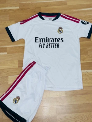 Real Madrid fotbollskit vit - Säljer ett vitt Real Madrid fotbollskit med matchande tröja och shorts. Tröjan har svarta detaljer vid ärmslut och krage, röda ränder på axlarna och klubbmärke på bröstet. Shortsen har röda ränder på sidan och klubbmärke. Perfekt för fotbollsträning.
