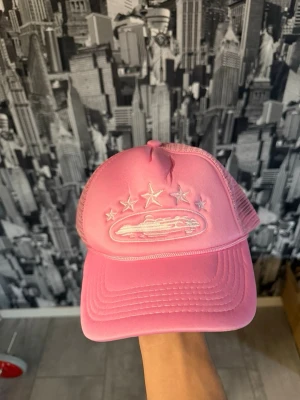 Rosa meshkeps från Corteiz - Säljer en rosa keps från Corteiz med mesh på sidorna och bak, justerbar snapback och broderad UFO och stjärnor framtill. Bak finns Corteiz-loggan broderad i vitt. Perfekt för dig som vill sticka ut med en unik och trendig accessoar.