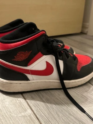 Air Jordan 1 Mid svart/röd/vit - Säljer ett par Air Jordan 1 Mid sneakers i svart, rött och vitt läder. Klassisk design med röd swoosh, svarta snören och ikonisk Jordan Wings-logga på sidan. Perfekta för dig som gillar streetwear och vill sticka ut med en tidlös modell.