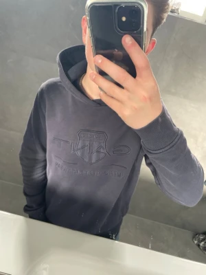 Mörkblå hoodie från GANT - Säljer en stilren mörkblå hoodie från GANT med broderad logga på bröstet. Hoodien har huva och ribbade muddar vid ärmslut och nederkant. Perfekt för en avslappnad och snygg look.
