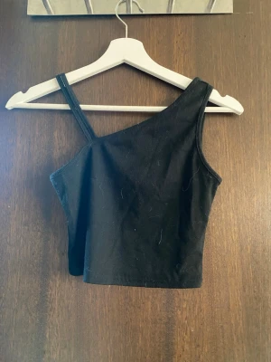 Svart oneshoulder croppad topp - Säljer en svart croppad topp med oneshoulder-design och ett smalt axelband. Toppens material är ribbad bomull vilket ger en snygg struktur och skön känsla. Perfekt för dig som gillar trendiga och stilrena plagg.
