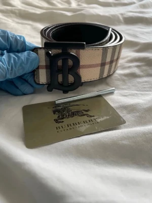 Burberry bälte med klassiskt rutmönster - Snyggt Burberry bälte med den ikoniska stora B-loggan i svart metall. Ena sidan har klassiskt beige, brunt och rött rutmönster, andra sidan är helsvart med diskret präglat mönster. Perfekt accessoar för att lyfta din outfit.