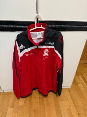 Röd och svart Adidas träningsjacka - Säljer en snygg röd och svart träningsjacka från Adidas med vita detaljer och dragkedja. Jackan har klubbtryck på bröstet, SpeediCath-logga och Barbro Sport-tryck. Perfekt för träning eller uppvärmning, med hög krage och långa ärmar.