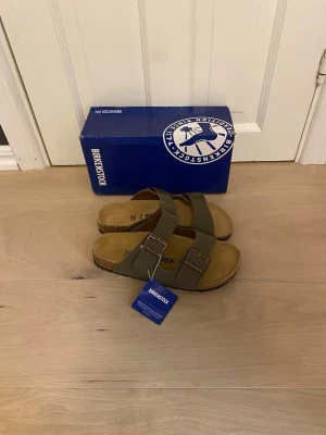 Birkenstock Arizona  - Säljer ett par olivgröna Birkenstock Arizona sandaler med två justerbara remmar och metallspännen. Fotbädden är i mocka och sulan är i kork för extra komfort. Klassisk och tidlös design som passar till det mesta.