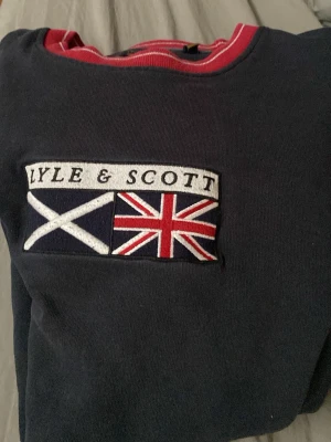 Mörkblå sweatshirt från Lyle & Scott - Mörkblå sweatshirt från Lyle & Scott med broderade flaggor på bröstet och kontrasterande röd krage. Tröjan har en klassisk rund halsringning och är tillverkad i mjukt material för en avslappnad look.