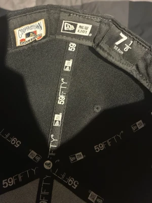 Svart New Era 59FIFTY keps 7 1/8 - Säljer en svart New Era 59FIFTY keps från Cooperstown Collection. Kepsen har en klassisk flat brim och är i storlek 7 1/8 (56.8 cm). Insidan är fodrad med märkets logga och etiketter. Perfekt för dig som gillar streetwear och sportig stil.