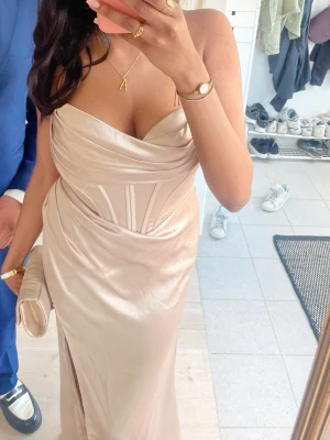 Champagnefärgad långklänning i satin - Elegant långklänning i champagnefärgad satin med draperad urringning och markerad midja. Klänningen har smala axelband och en slits framtill för en stilren look. Perfekt för speciella tillfällen.