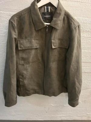 Olivgrön overshirt från Dressmann mocca - Stilren olivgrön overshirt från Dressmann i mjukt mockaimitationsmaterial. Jackan har två stora bröstfickor med lock, dragkedja framtill och klassisk krage. Perfekt lager på lager-plagg för en avslappnad look. Mocca