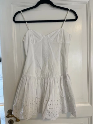 Vit kort klänning med broderad spets Zara - Söt vit kort klänning med tunna axelband och broderad spetsdetalj längst ner. Klänningen har en feminin passform och är perfekt för varma dagar. Tillverkad i bomull som känns lätt och skönt mot huden. Har en underklänning. Passar dig som gillar en enkel och fräsch stil. Perfekt för student eller en härlig sommardag.