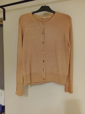 Beige cardigan med gulddetaljer (Kofta, NY) - Stilren beige cardigan med rund halsringning och guldfärgade knappar framtill samt på ärmsluten. Tunnstickad modell med ribbade muddar och slits vid ärmslut. Perfekt att slänga över en topp för en clean look. Super fin kofta helt ny!