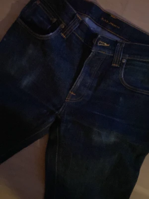 Mörkblå jeans från Nudie Jeans - Snygga mörkblå jeans från Nudie Jeans med klassisk femficksdesign och raka ben. Jeansen har kontrastsömmar och är tillverkade i bomull. Perfekta för en stilren och avslappnad look. Storlek 31/32