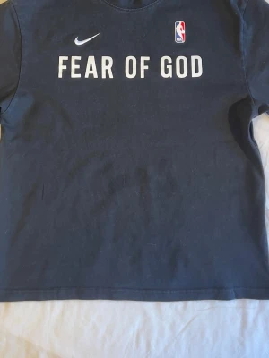 Svart Fear of God x Nike NBA t-shirt - Säljer en svart t-shirt från Nike i samarbete med Fear of God och NBA. T-shirten har tryck framtill med texten 'FEAR OF GOD', Nike-logga och NBA-logga. Klassisk passform och rund halsringning. Perfekt för dig som gillar streetwear och basket.