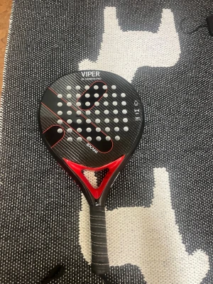 Viper X Carbon Pro padelracket - Säljer ett Viper X Carbon Pro padelracket i svart och rött med kolfiberyta och perforerat mönster. Racket har greppvänligt handtag med vit söm och modern design. Perfekt för dig som gillar padel och vill ha ett snyggt och stabilt racket. Inte använt det så mycket då padel inte var min grej.