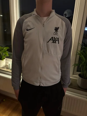 Nike LFC träningsjacka vit/grå - Snygg träningsjacka från Nike i vitt med grå ärmar och svarta detaljer. Jackan har dragkedja, LFC-logga och AXA-tryck på bröstet. Perfekt för dig som gillar fotboll och vill ha en sportig look. Materialet är mjukt och följsamt. passar dig som är runt 170