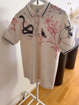 Vit kortärmad skjorta med tryck Zara - Vit polo t-shirt från Zara med unikt tryck av ormar, blommor och insekter i svart Tshirten har krage med knappar och svarta ränder på kragen och ärmsluten. Perfekt för dig som vill sticka ut med en cool och konstnärlig stil. Storlek L 