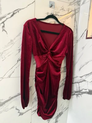 Vinröd sammetsklänning med v-ringning - Vinröd kort sammetsklänning med lång ärm och v-ringning. Klänningen har en snygg twist-detalj framtill och rynkade sidor som ger en smickrande passform. Inga skador eller defekter 