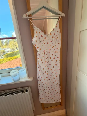 Vit blommig långklänning med smala axelband - Superfin vit långklänning med färgglatt blommönster och smala axelband. Klänningen har v-ringning och en lätt figurnära passform, perfekt för varma dagar. Materialet känns svalt och mjukt mot huden.