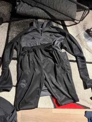 Svart Real Madrid träningsset Adidas M-L - Säljer ett svart träningsset från Adidas och Real Madrid. Setet består av en långärmad tröja med halv dragkedja och matchande byxor, båda med Real Madrid-logga och Adidas-märke. Materialet är mjukt och stretchigt, perfekt för träning eller uppvärmning.
