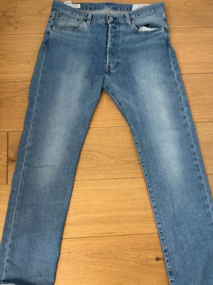 Levi's 501  - Säljer ett par klassiska ljusblå jeans från Levi's med rak passform och fem fickor. Jeansen har knappgylf och subtila slitningar för en avslappnad look. Tillverkade i bomullsdenim och har den ikoniska läderlappen bak i midjan.