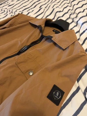 Overshirt  - Snygg brun overshirt från Lagency med dragkedja framtill och bröstficka med knapp. Patch med logotyp på ärmen ger en cool detalj. Materialet är lätt och passar perfekt för lager-på-lager. Klassisk krage och långärmad modell. Endast använd enstaka tillfällen.