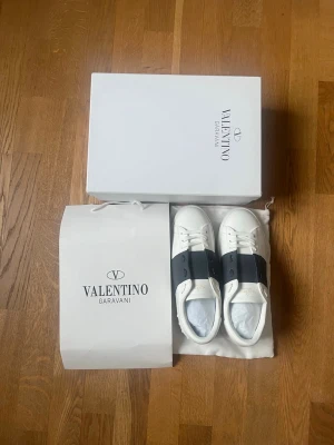 Valentino Garavani vita sneakers - Snygga vita sneakers från Valentino Garavani med bred svart läderdetalj över snörningen. Klassisk låg modell med rund tå och vita skosnören. Perfekt för dig som vill ha en exklusiv och stilren look.