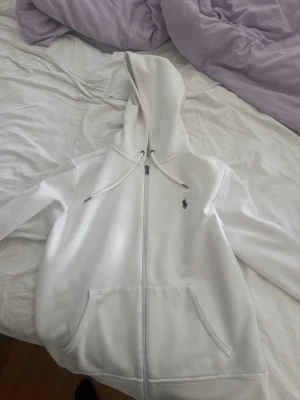 Vit hoodie från Polo Ralph Lauren - Säljer en clean vit hoodie från Polo Ralph Lauren med dragkedja, huva och fickor framtill. Klassisk logga broderad på bröstet. Perfekt för dig som gillar stilrena plagg med premiumkänsla. Materialet är mjukt och skönt, passar till alla tillfällen.