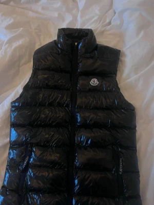 Svart dunväst från Moncler - Snygg svart dunväst från Moncler med glansig finish och klassisk logga på bröstet. Västen har hög krage, dragkedja framtill och två fickor med dragkedja. Perfekt för lager-på-lager och streetstyle.