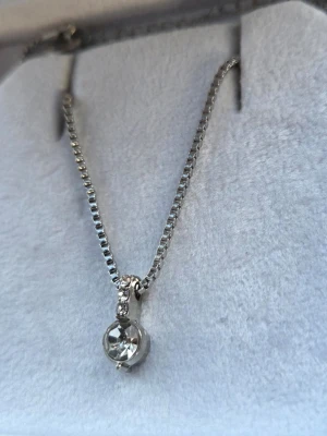 Silverfärgat halsband med glittrande sten - Snyggt halsband i silverfärg med en glittrande rund klar sten som hänge. Kedjan är boxlänk och hänget har små gnistrande stenar ovanför den stora stenen. Perfekt accessoar för att lyfta din outfit.