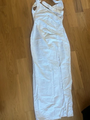 Vit långklänning med slits - Snygg vit långklänning med omlott-detalj och slits framtill. Klänningen har ärmlös design och smala axelband, vilket ger en stilren och modern look. Perfekt för varma dagar eller speciella tillfällen.
