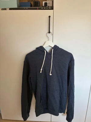 Mörkblå hoodie med vit dragsko - En stilren mörkblå hoodie med huva och vita dragsnören, knappt använd från pier one. Hoodien har en klassisk känguruficka framtill och är tillverkad i mjukt material som känns skönt mot huden. Perfekt för en avslappnad look och enkel att matcha med jeans eller joggers.