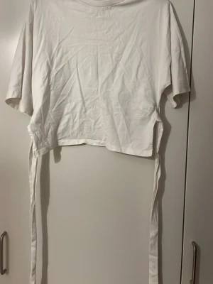 Vit topp med knyt från Nelly - Behövs strykas!! Vit kortärmad topp från Nelly med bred ribbad krage och långa knytband i sidorna. T-shirten har en loose passform och är tillverkad i mjuk bomull, perfekt för en avslappnad stil. Köp så snabbt som möjligt!💕