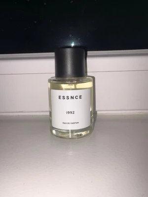 ESSNCE 1992 Eau de Parfum - Parfymflaska från ESSNCE med namnet 1992. Flaskan är genomskinlig med en vit etikett och svart text, och har ett svart cylinderformat lock. Innehållet är ljust gult. Perfekt för dig som gillar stilren design. Säljer den för de inte va min smak av parfym, har även inte använt den så den är 50 ml.