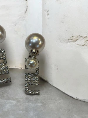  örhängen med pärlor - Statementörhängen med stora silvriga pärlor och gnistrande kristallbokstäver. Bokstäverna är täckta av små stenar som ger extra bling och örhängena har en lyxig och trendig vibe. Perfekta för dig som vill sticka ut och visa din stil.
