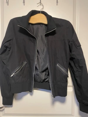 Svart bomberjacka med dragkedjor - Snygg svart bomberjacka med silvriga dragkedjor och ribbade muddar. Jackan har två snedställda fickor framtill. Klassisk bombermodell med hög krage och fodrad insida. Perfekt för en streetwear-look.